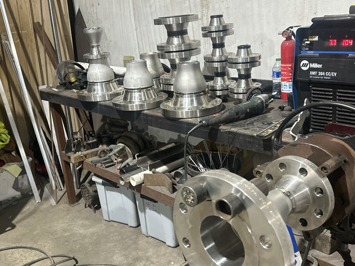 Batch of stainless steel reducers and flanges on a workbench next to a Miller welder